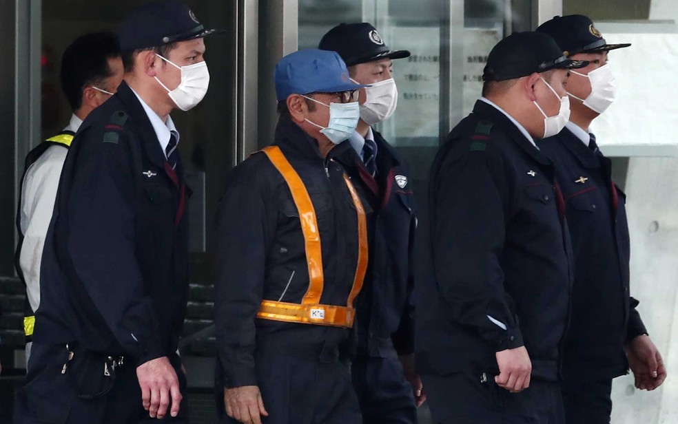 Brasileiro Carlos Ghosn (ao centro, de boné azul e máscara) é escoltado ao sair da Casa de Detenção de Tóquio — Foto: Behrouz Mehri / AFP Photo